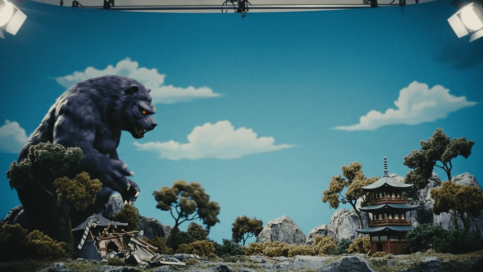 A dynamic cinematic styled like a 1970s Japanese tokusatsu battle scene. Two giant rubber-suit monsters clash in the middle of a destroyed miniature city set. a giant purple monster walks into the movie set each step shakes the scene the monster poses angry The environment is a detailed miniature downtown: crumbling model buildings, scattered rubble, toy cars, smoke plumes, and broken street lamps. The scale feels large but clearly practical and handmade. Shot from a slightly low Dutch angle to emphasize size and heroism. The background features a dramatic painted sunset sky with stylized clouds in purples, oranges, and deep blues. Lighting is strong and theatrical, with studio spotlights creating dramatic highlights and deep shadows across the rubber suits. The overall look mimics vintage practical effects: visible seams, suit wrinkles, matte textures, chunky miniatures, and practical fire/smoke elements. Captured with a retro 35mm tokusatsu aesthetic: vivid saturated colors, slight grain, mild softness at the edges, and the charming imperfection of old special-effects shows. The moment shows both monsters mid-fight, poised for impact, full of energy and vintage cinematic flair