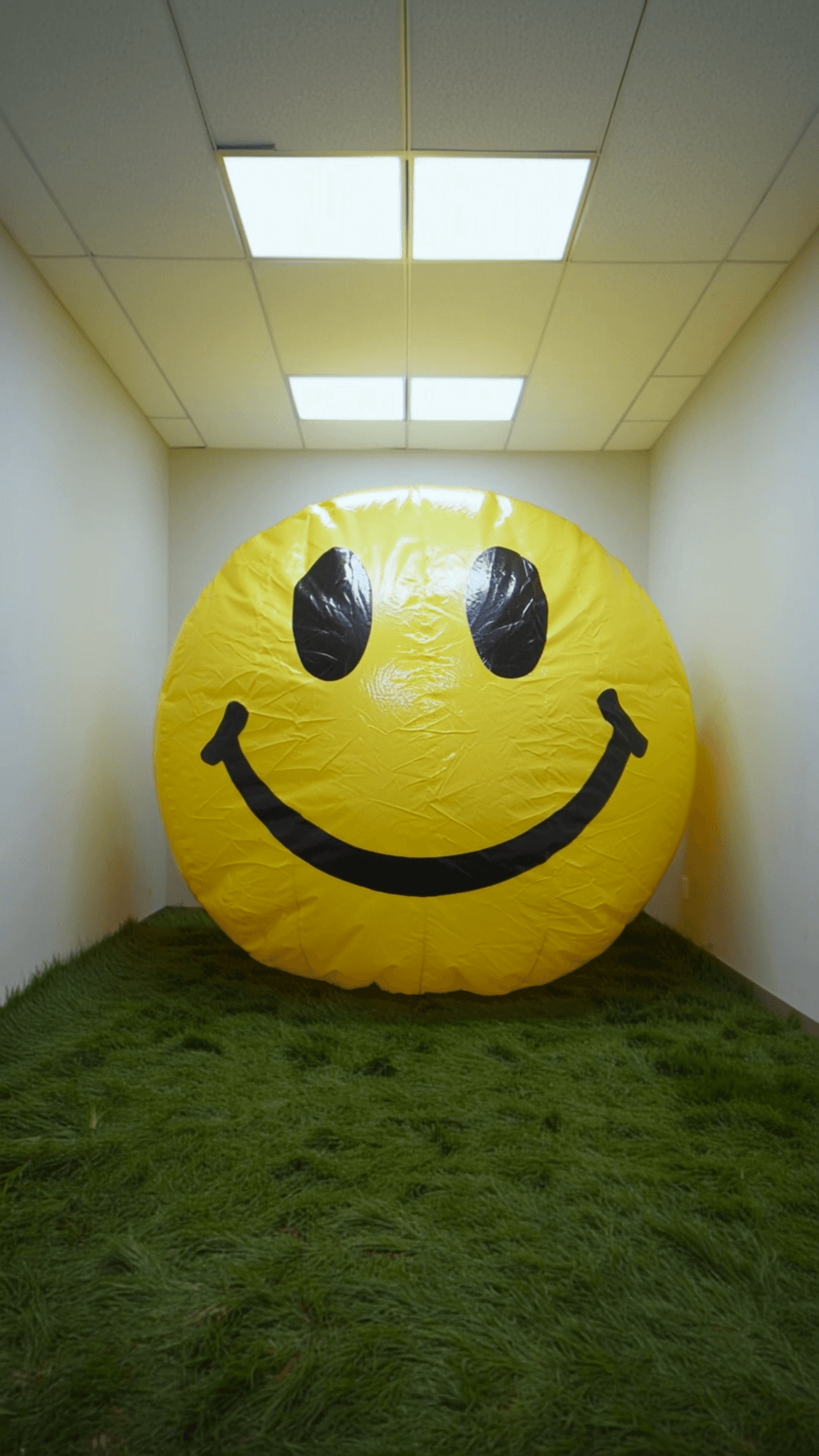 Ultra-realistic cinematic wide-angle shot inside an empty 1980s corporate office interior. No furniture. No objects. Only plain off-white walls a low ceiling with rectangular fluorescent light panels clearly indoors. The entire floor is covered in real natural grass thick and uneven clearly growing indoors. At the start of the shot a large but not fully inflated inflatable smiley face sits several feet away from the camera. The inflatable is neon yellow glossy rubber-like with visible seams folds and wrinkles. It has simple black oval eyes and a thick curved black smile. Over time the inflatable continuously inflates without stopping. As it inflates it expands toward the camera in addition to pressing into walls ceiling and floor. The grass is flattened beneath it. The inflatable deforms heavily under pressure stretching and bulging. By the end of the shot the inflatable fills the entire room volume AND pushes directly into the camera lens. The smiley face makes physical contact with the lens occupying the entire frame. Perspective distorts as the surface presses forward. The final frame is extreme close-up with the inflatable touching the lens causing slight warping and softness at the edges. Lighting remains cold flat fluorescent with bright specular highlights sliding across the surface during contact. Camera is static wide-angle (18–22mm) and does not move or pull back. The camera cannot escape the inflation. Photorealistic materials global illumination cinematic realism. Overwhelming claustrophobic unavoidable. No people. No logos. No branding. 
