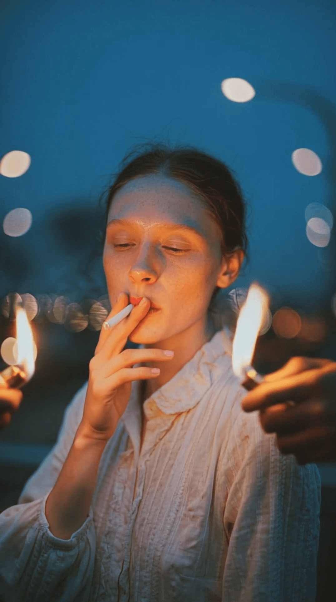 ultra-cinematic night portrait of a woman in the center of the frame intense focused expression looking down as she lights a cigarette dramatic close-up composition surrounded by many hands reaching in from all sides holding lit lighters at least seven hands total eight lighters total flames creating warm highlights on her face and shoulders deep blue twilight sky background subtle city lights bokeh far in the distance high contrast lighting moody film still shallow depth of field sharp realistic skin texture glossy reflections on metal lighters hands layered in foreground creating depth 35mm cinematic look rich color grading gritty editorial vibe natural film grain crisp focus dramatic rim light and flame glow realistic smoke haze immersive intimate framing. she putts a n un litted cigareete in her mouth and the hands enter the frame to light the cigarette